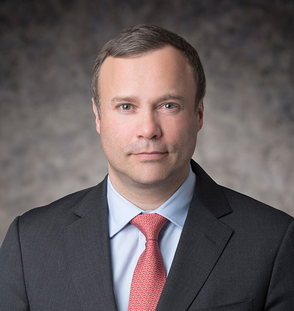 Dr Remy Gerbay in a grey suit, red tie and blue shirt against a grey bacground