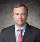 Dr Remy Gerbay in a grey suit, red tie and blue shirt against a grey bacground