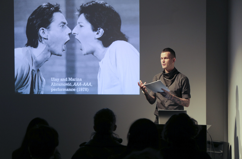 Dominic Johnson gives a lecture backed by a slide showing a performance by Ulay/Abramovic