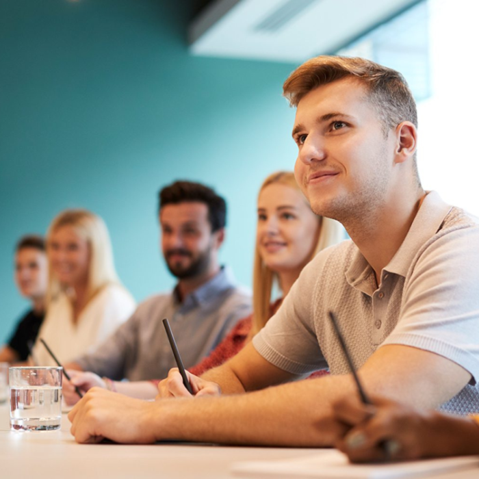 Various students sitting at a table holding pens and listening to a tutor