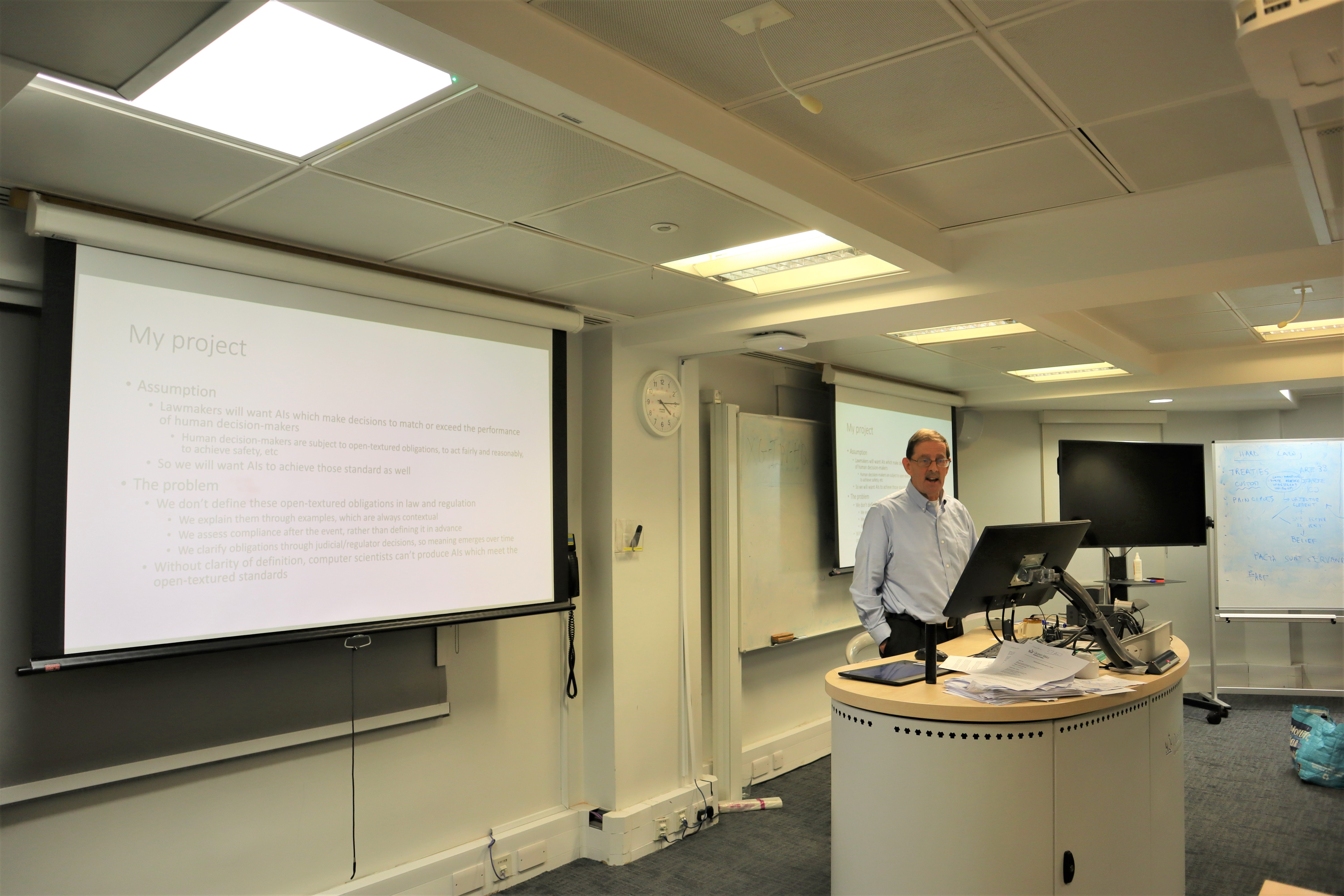 Professor stood at lecturn with projector displaying slides behind