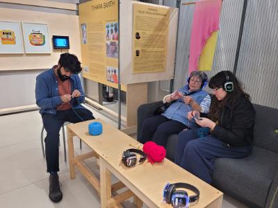 Gallery visitors listening to part of the exhibition on headphones whilst knitting.