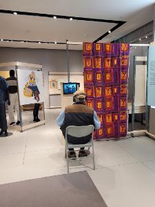 A man sat in a chair wearing headphone and watching a video. He is next to a red, yellow, and orange textile curtain.