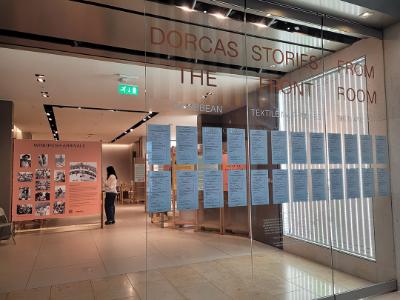 A glass door with a sign for the Dorcas Stories exhibition. Behind it is an information board on Windrush Arrivals.