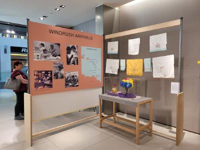 An information board about Windrush Arrivals. Next to it are some textile artwork and sculptures.