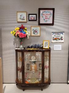 A glass cabinet with artwork displayed inside. On top is a vase full of flowers and some hanging pictures.