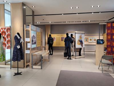 People walking through an the Dorcas exhibition. There are panels about the artists and some textiles on display including a navy and white dress on a mannequin.