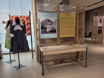 A panel on Windrush Futures. Next to the panel are mannequins. One is dressed in a navy dress with white patterns. The other is a white dress with a light green fern pattern.