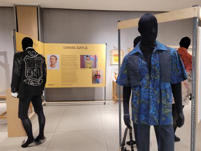  A panel on Daniel Gayle. There are mannequins. One is wearing a blue shirt with a flowing water pattern and one is wearing a dark denim jacket with a modern coat of arms design.