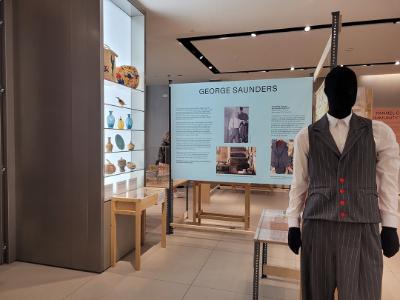 A mannequin dressed in a navy pinstripe suit. Behind is an information board for George Saunders and a shelf of decorated vases and pottery.