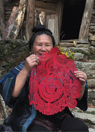 A woman from Guizhou practising Miao paper-cutting. She is holding up the deep red paper and smiling.