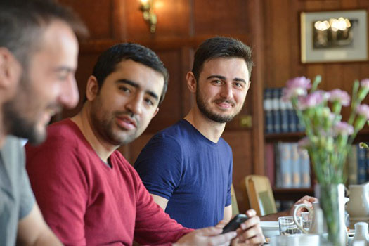 Three students sat in a line having a discussion. One is looking to the camera and smiling.
