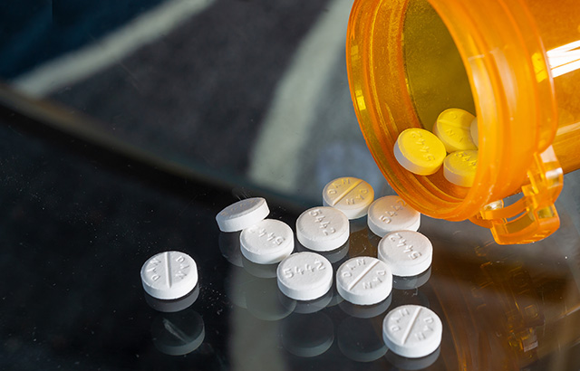 An open orange pill bottle tipped over on to a table with white tablets spilling out.