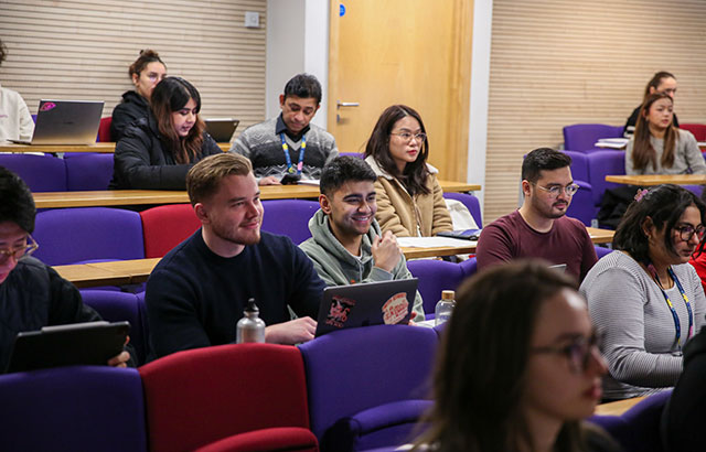 Group of students sit in a lecture theatre on a mixture of coloured chairs