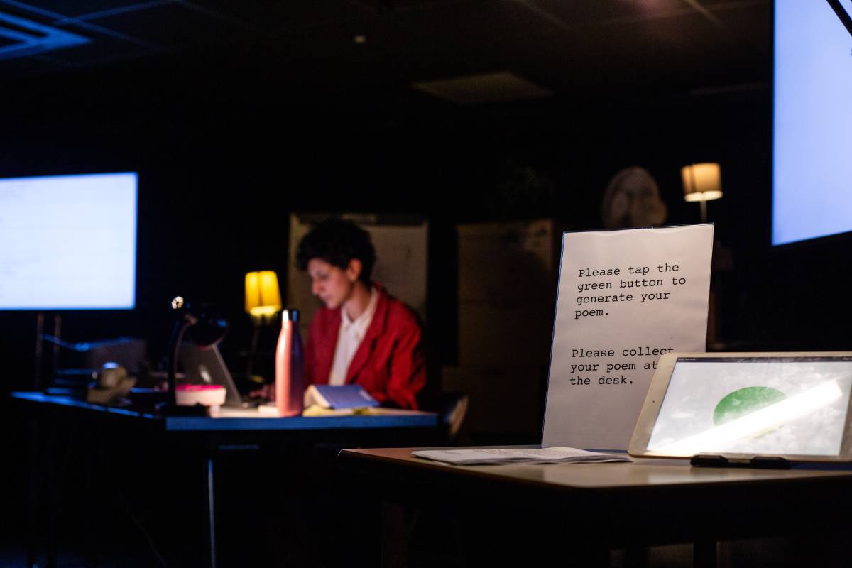 A poet sits at a desk with a low lit lamp, behind a sign that reads 'please tap the green button to generate your poem'