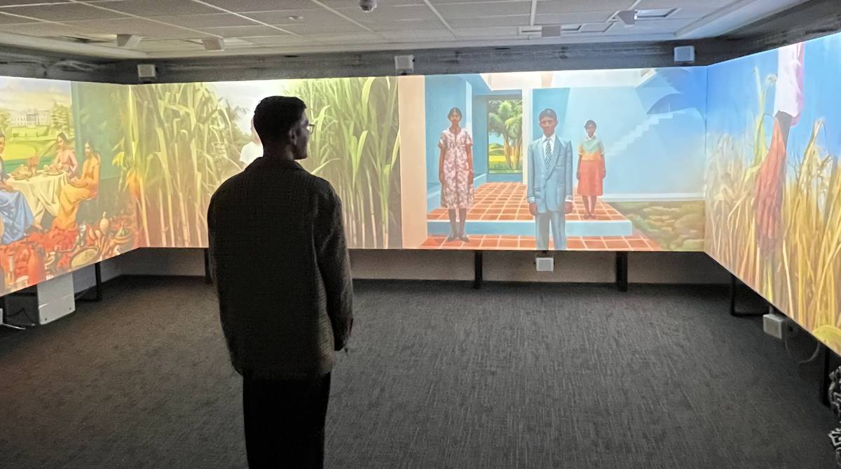 A man stands in front of a colourful projection of artwork depicting South Asian family life
