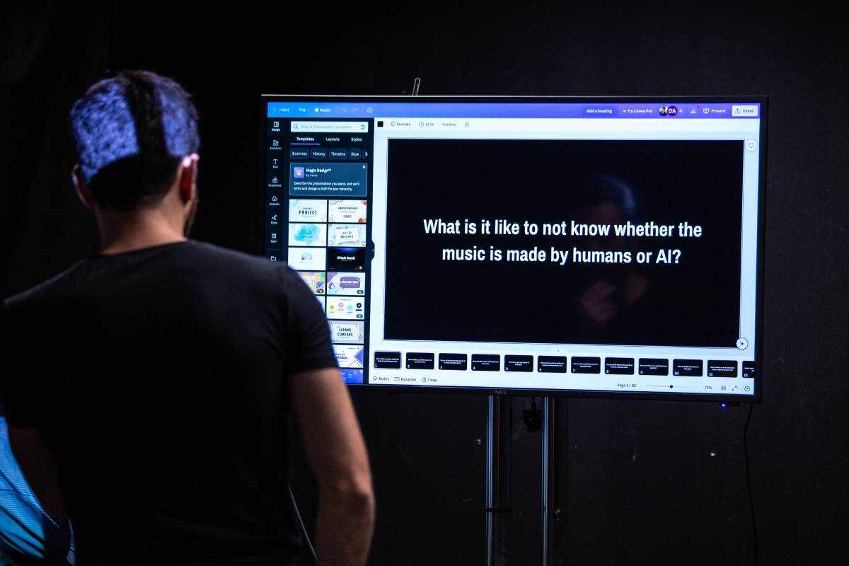 A man stands in front of several screens, reading the words 'what is it like to not know if the music is made by humans or AI?'