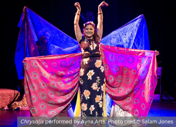 On stage a smiling woman stands with her hands in the air, with colourful silks pulled out from behind her like butterfly wings. From the show 
