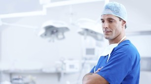 A doctor standing with cross armed in a theatre wearing scrubs and a sergical hat