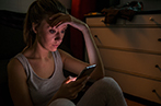 A woman sat on the floor of her room in the dark looking worriedly at her phone.