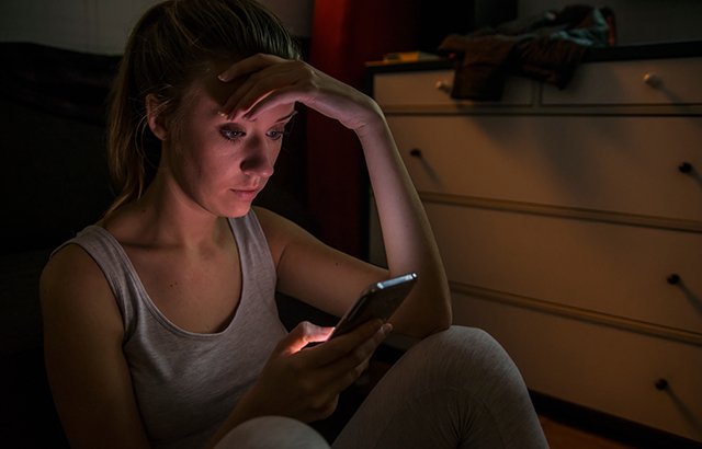 A woman sat on the floor of her room in the dark looking worriedly at her phone.