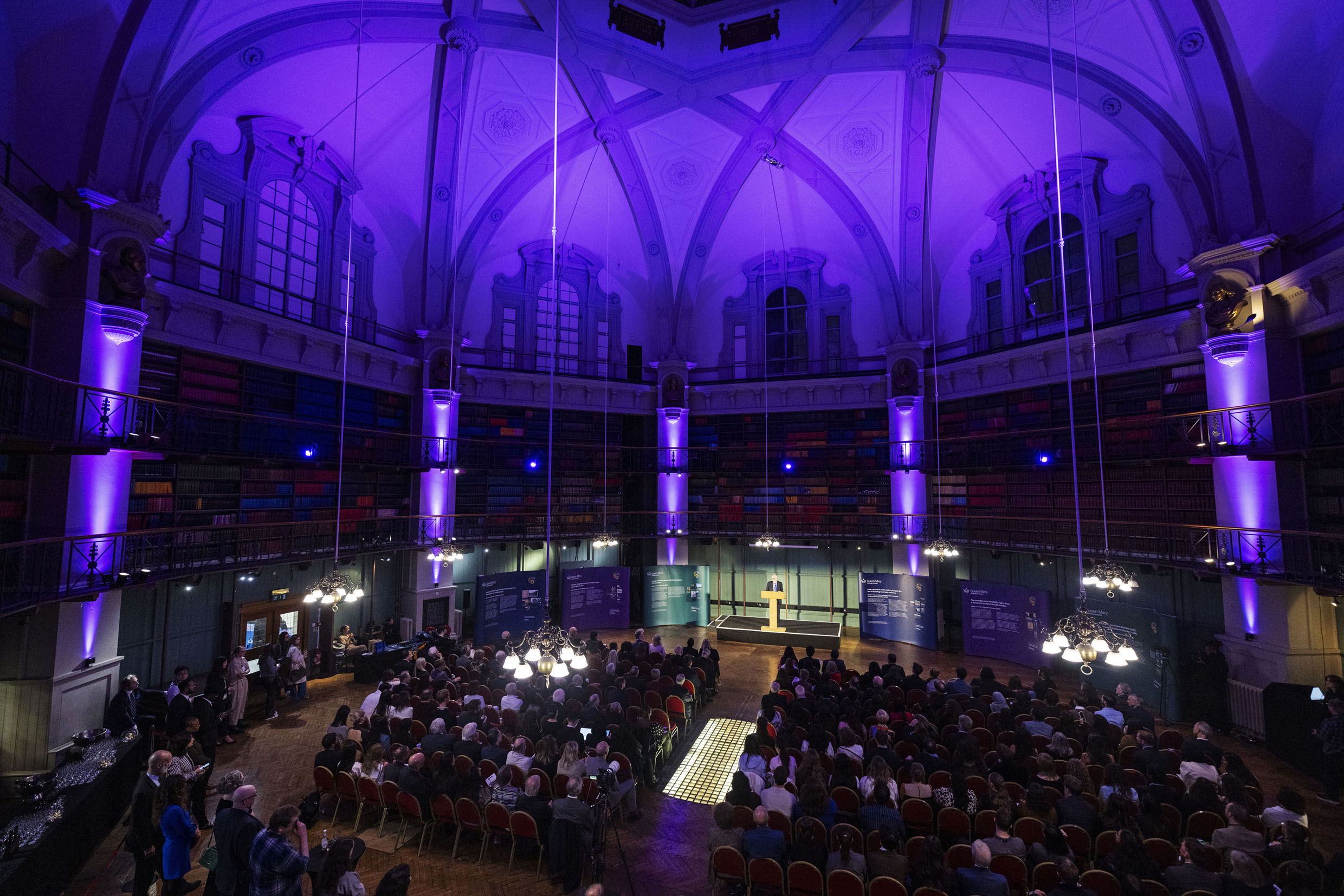 Octagon room lit in purple filled with an audience listening to a speaker on stage.