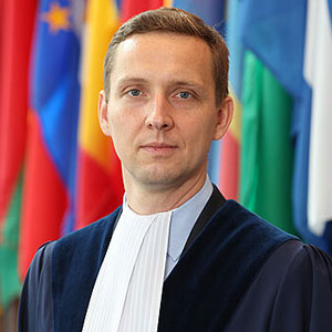 Judge Konrad Jan Marciniak wearing his gown standing in front of various flags.