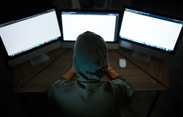 Person wearing a hoodie, with the hood up, looking at computer monitors.