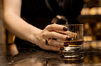 A person, presumably a woman, with their nails painted black and drinking a glass of whiskey in a bar.