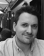 Manuel Gonzalez. The photo is in black and white. He is sitting outside a café, smiling.