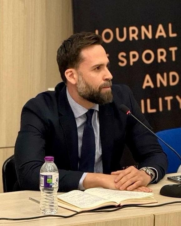 Nick wears a dark suit and sits at a table with a microphone with a notebook in front of him