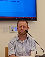 Dr Noam Gur speaking at an event. He is sat down, wearing a blue shirt and at a microphone.