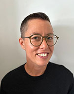 Sarah Keenan. They are standing against a white wall wearing glasses. They are smiling.