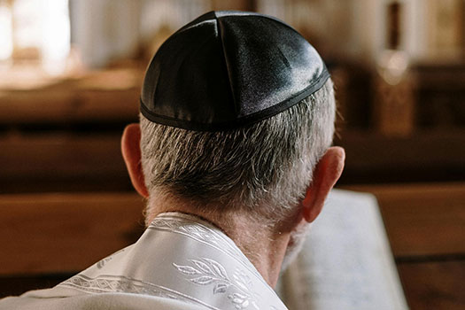 View of the back of a man's head. He is sat in a synagogue wearing a kippa.