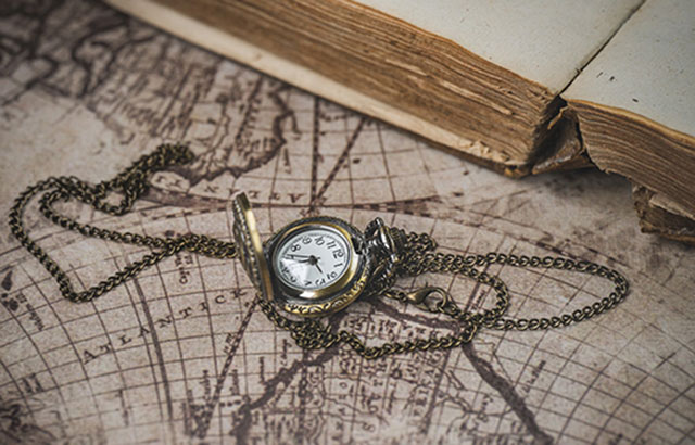 A vintage pocket watch laid on top of an old map. An old book is laid open at the top of the frame.