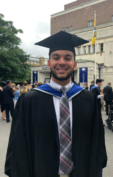Paul stands squarely facing the camera in graduation cap and gown. He is also wearing a white collared shirt and a tie. He has olive skin and close-cut beard. He has a wide, bright smile.