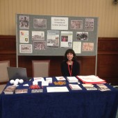 Colour image of a woman sitting behind a table; there is a noticeboard behind them decorated with photographs and leaflets on the table