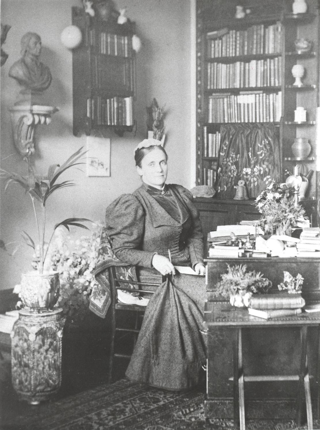Photograph of Constance Maynard seated at her desk in her office looking at the camera and holding a pen