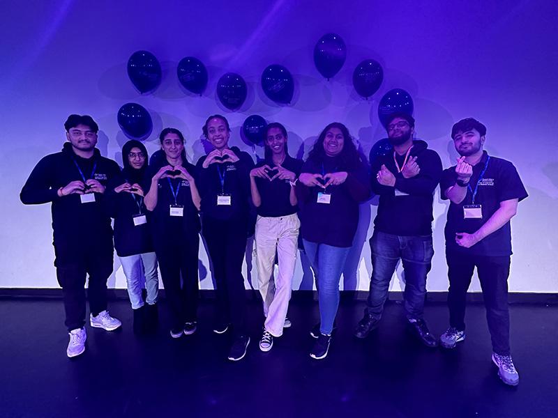 Student ambassadors making a heart sign with their hands, there are balloons behind them