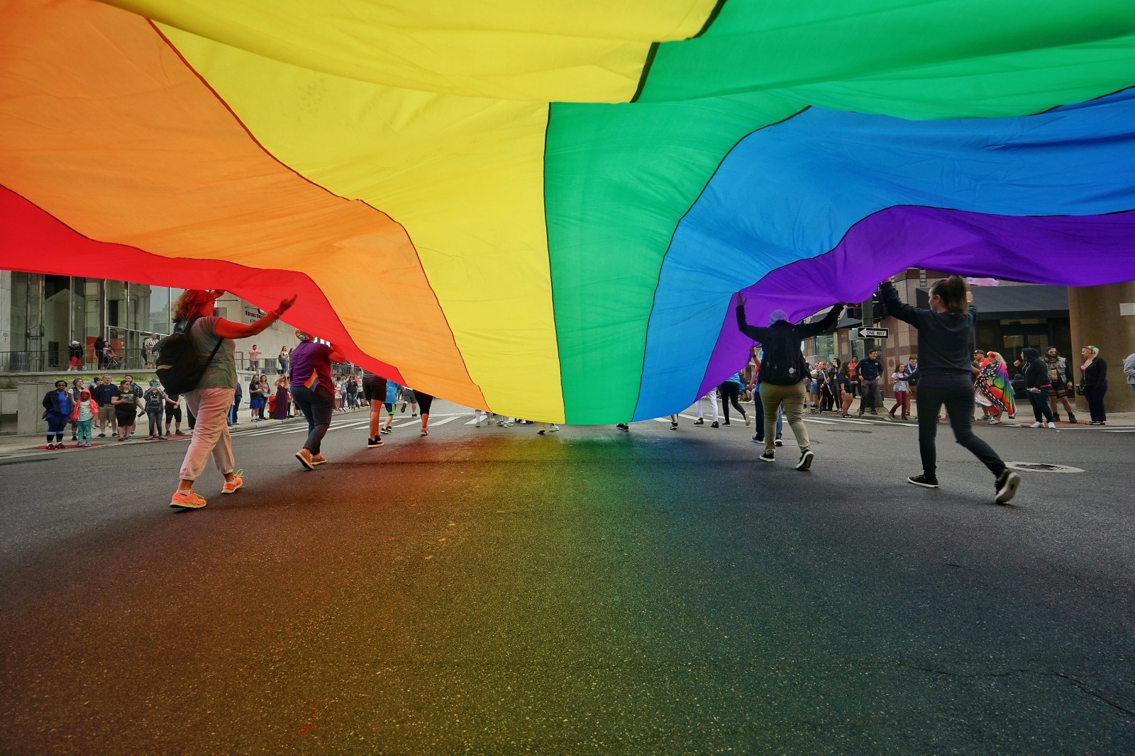 A large rainbow flag is being carried overhead by a group of people walking down a city street during a parade. The fabric creates a colourful canopy in red, orange, yellow, green, blue and purple, with participants visible beneath it and spectators lining the pavement in the background. The scene is lively and celebratory, capturing movement, community, and pride.