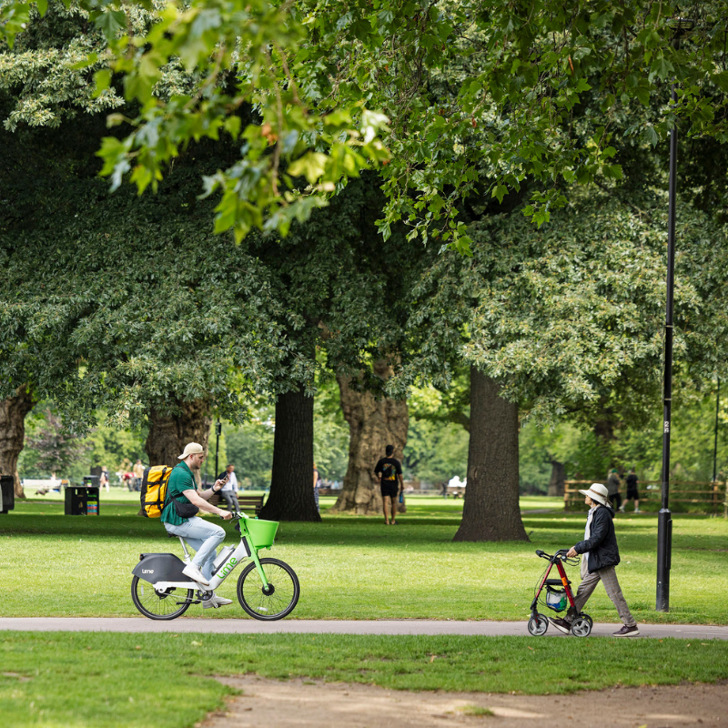 In a green park, someone cycles and another walks with a mobility aid