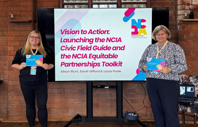 Image of two Queen Mary colleagues, each holding a booklet in their hands and one standing on either side of a TV screen, with a brick wall behind them.