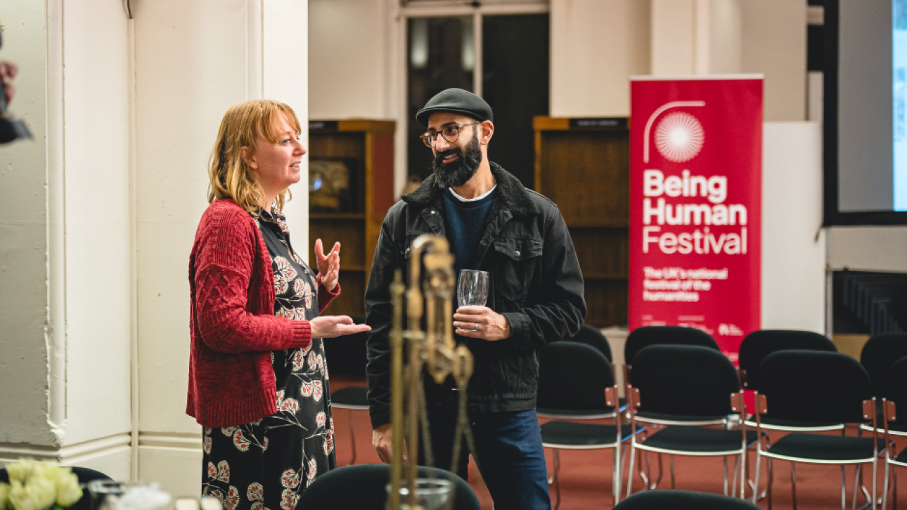 Two people stand in front of a Being Human Festival banner talking