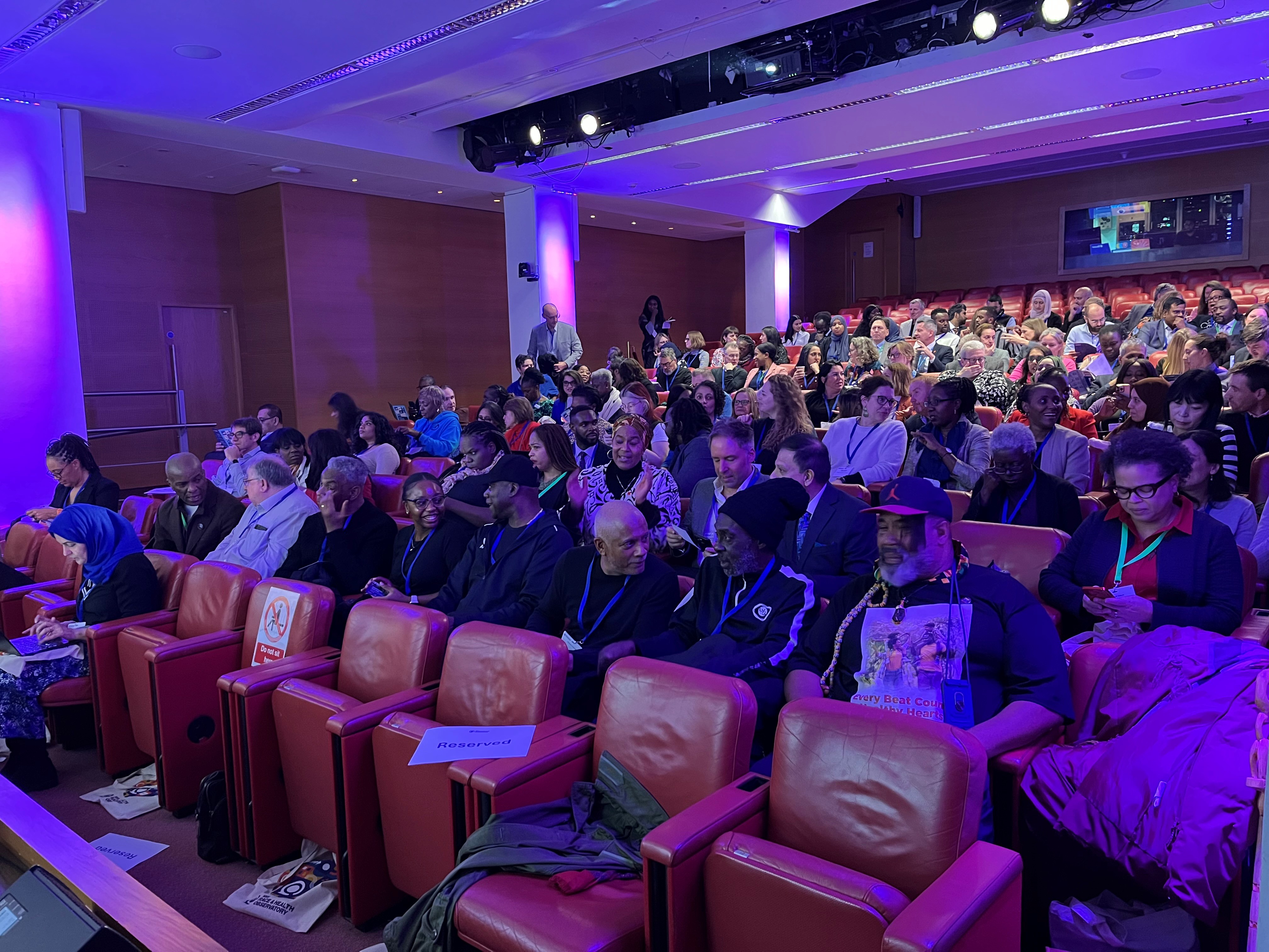 A crowd of people sit in cinema style seating in a purple lit room