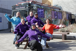 A group of adults cheer in front of a bus painted like the night sky named 
