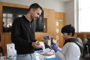 A child wearing goggles and holding a vial of red liquid speaks with an adult