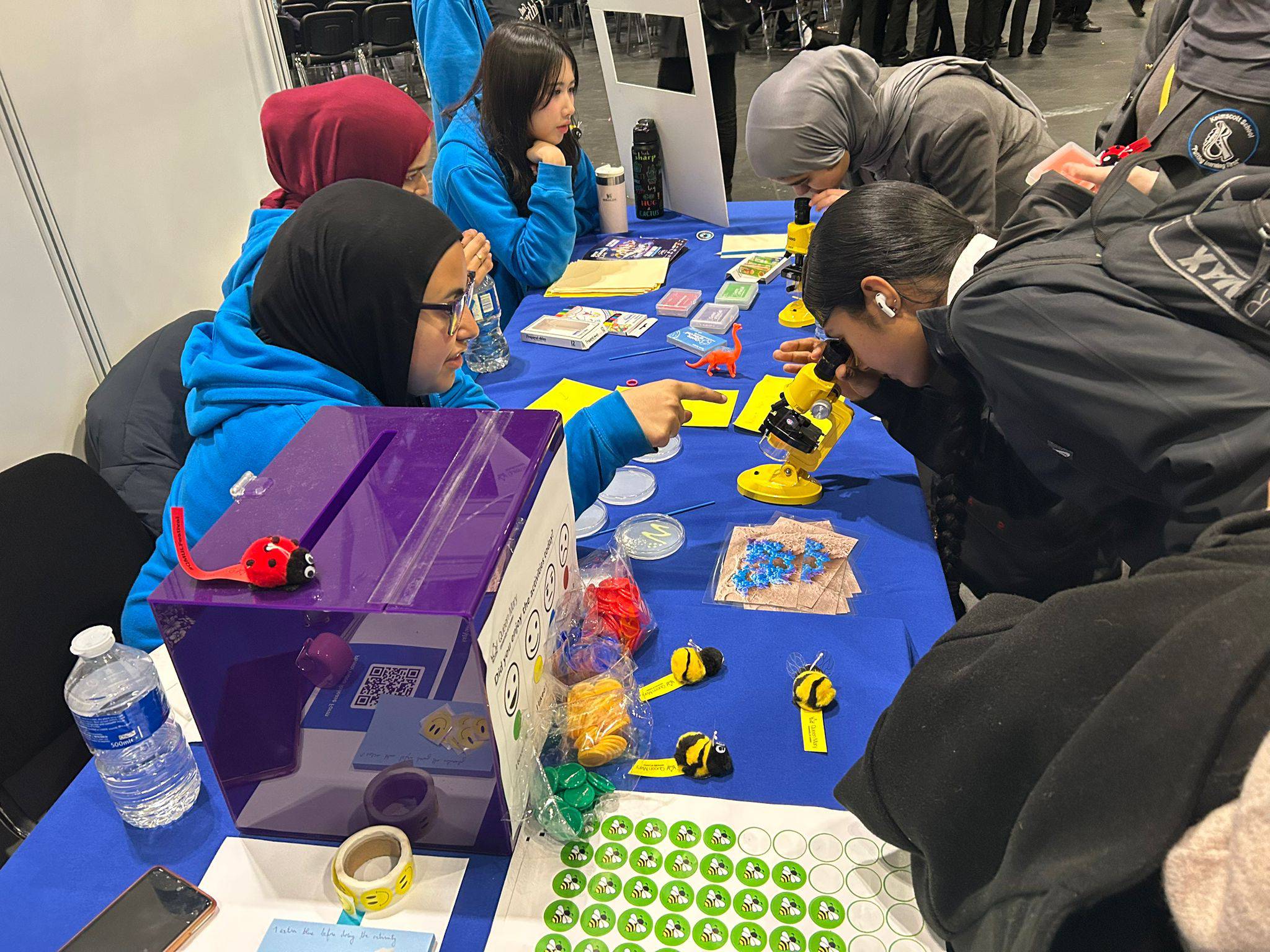 At a table with a blue tablecloth, university students show secondary school students how to use a microscope