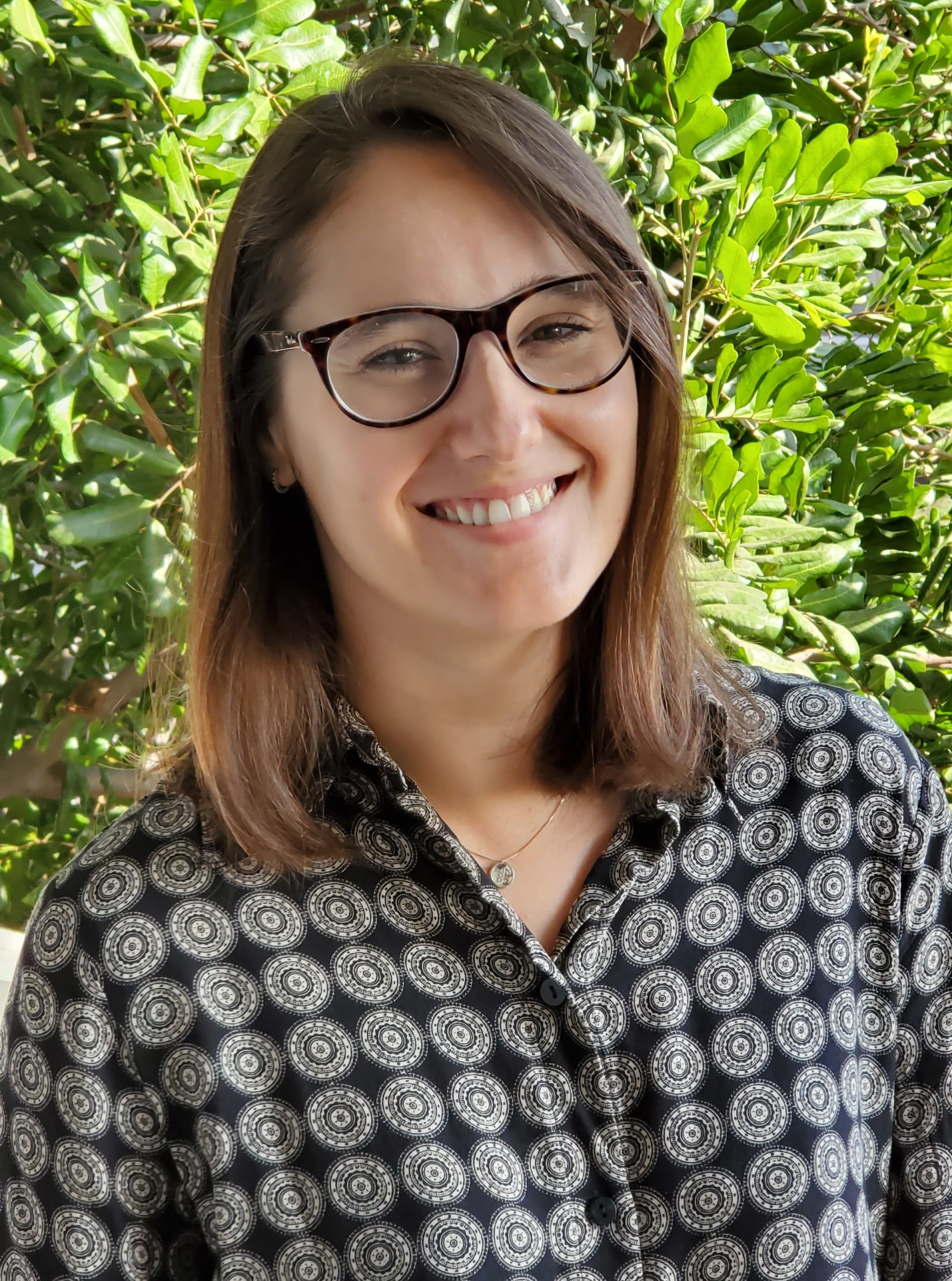 A headshot of a person with brown glasses and brown hair in front of foliage