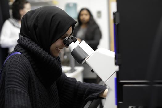 A woman in a hijab looking into a microscope smiling