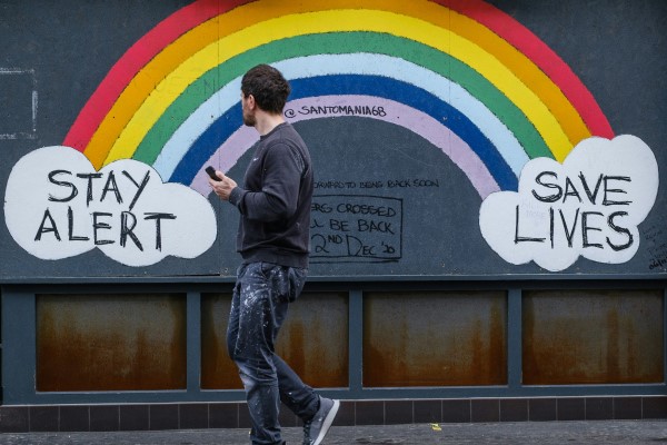 A photo of a person walking in front of a rainbow mural. At one end there is a cloud with text saying 'stay alert' and at the other, text saying 'save lives'.
